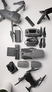 A collection of aerial drone equipment is neatly arranged on a white surface. This includes two drones with propellers, a remote control, multiple batteries, a camera, a set of propeller blades, charging cables, and a tripod. The items are predominantly gray and black, giving a sleek, professional appearance.