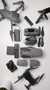 A collection of aerial drone equipment is neatly arranged on a white surface. This includes two drones with propellers, a remote control, multiple batteries, a camera, a set of propeller blades, charging cables, and a tripod. The items are predominantly gray and black, giving a sleek, professional appearance.