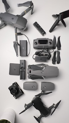 A collection of aerial drone equipment is neatly arranged on a white surface. This includes two drones with propellers, a remote control, multiple batteries, a camera, a set of propeller blades, charging cables, and a tripod. The items are predominantly gray and black, giving a sleek, professional appearance.