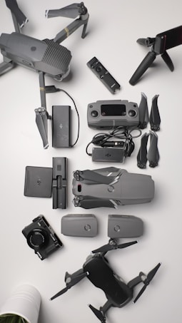 A collection of aerial drone equipment is neatly arranged on a white surface. This includes two drones with propellers, a remote control, multiple batteries, a camera, a set of propeller blades, charging cables, and a tripod. The items are predominantly gray and black, giving a sleek, professional appearance.