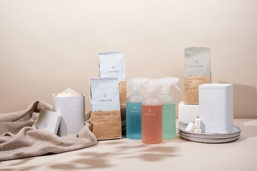 A minimalist arrangement of eco-friendly cleaning products including various bottles, containers, and packaging. The items are neatly placed on a soft beige fabric backdrop, emphasizing a sustainable and natural aesthetic.