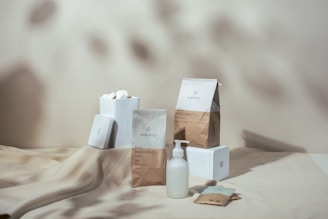 Aesthetic display of items including a dispenser bottle, paper packs, and white boxes placed on a light-colored surface with soft shadows in the background. The items feature minimalist designs and branding.