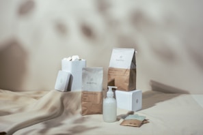 Aesthetic display of items including a dispenser bottle, paper packs, and white boxes placed on a light-colored surface with soft shadows in the background. The items feature minimalist designs and branding.