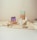 A minimalist composition featuring eco-friendly cleaning products arranged on a textured beige fabric. There's a bottle with a pump dispenser, a small packet, and two paper bags with lavender and mint green labels. The background is softly lit, creating a calm atmosphere.