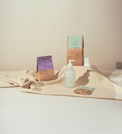 A minimalist composition featuring eco-friendly cleaning products arranged on a textured beige fabric. There's a bottle with a pump dispenser, a small packet, and two paper bags with lavender and mint green labels. The background is softly lit, creating a calm atmosphere.