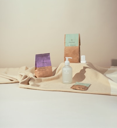 A minimalist composition featuring eco-friendly cleaning products arranged on a textured beige fabric. There's a bottle with a pump dispenser, a small packet, and two paper bags with lavender and mint green labels. The background is softly lit, creating a calm atmosphere.