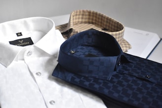 Three neatly folded dress shirts are placed on a flat surface. The shirts include a white one with a subtle pattern, a navy blue shirt with a checkered pattern, and a beige shirt with a plaid pattern. Each shirt is accompanied by paper packaging with brand labeling visible on the white shirt.