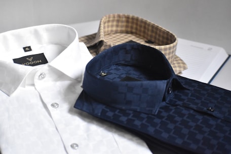 Three neatly folded dress shirts are placed on a flat surface. The shirts include a white one with a subtle pattern, a navy blue shirt with a checkered pattern, and a beige shirt with a plaid pattern. Each shirt is accompanied by paper packaging with brand labeling visible on the white shirt.