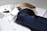 Three neatly folded dress shirts are placed on a flat surface. The shirts include a white one with a subtle pattern, a navy blue shirt with a checkered pattern, and a beige shirt with a plaid pattern. Each shirt is accompanied by paper packaging with brand labeling visible on the white shirt.