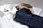 Three neatly folded dress shirts are placed on a flat surface. The shirts include a white one with a subtle pattern, a navy blue shirt with a checkered pattern, and a beige shirt with a plaid pattern. Each shirt is accompanied by paper packaging with brand labeling visible on the white shirt.