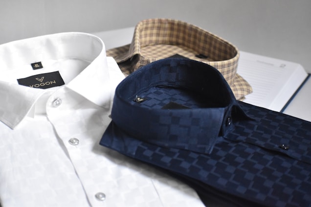 Three neatly folded dress shirts are placed on a flat surface. The shirts include a white one with a subtle pattern, a navy blue shirt with a checkered pattern, and a beige shirt with a plaid pattern. Each shirt is accompanied by paper packaging with brand labeling visible on the white shirt.