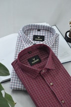 Two neatly folded collared shirts are placed on top of an open notebook. The shirts feature a checkered pattern, one predominantly red and the other predominantly blue. The brand label 'VOGON' is visible on both shirts.