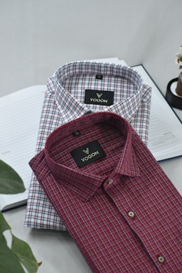 Two neatly folded collared shirts are placed on top of an open notebook. The shirts feature a checkered pattern, one predominantly red and the other predominantly blue. The brand label 'VOGON' is visible on both shirts.