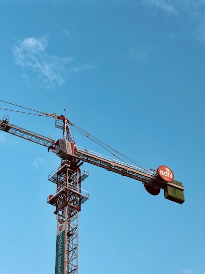 A large construction crane extends against a clear blue sky, with a circular sign displaying 'adhi' attached near the lifting mechanism. The crane's structure is detailed and industrial, showing various cables and metal frameworks.