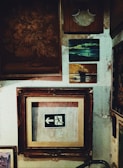 A collection of framed artworks and photographs is displayed on a textured wall. The centerpiece is a framed emergency exit sign with an arrow pointing left. Surrounding it are various other frames, including a painting of a ship at sea and an abstract floral piece. The wall surface appears worn and aged, adding a rustic ambiance.