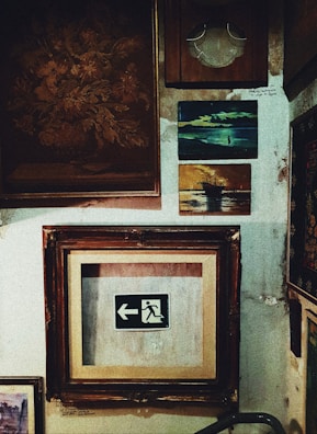 A collection of framed artworks and photographs is displayed on a textured wall. The centerpiece is a framed emergency exit sign with an arrow pointing left. Surrounding it are various other frames, including a painting of a ship at sea and an abstract floral piece. The wall surface appears worn and aged, adding a rustic ambiance.