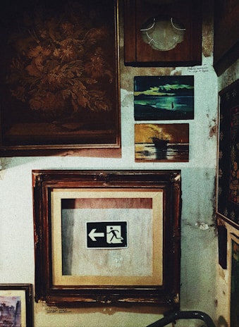 A collection of framed artworks and photographs is displayed on a textured wall. The centerpiece is a framed emergency exit sign with an arrow pointing left. Surrounding it are various other frames, including a painting of a ship at sea and an abstract floral piece. The wall surface appears worn and aged, adding a rustic ambiance.