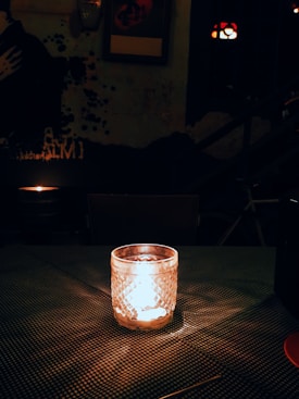 A dimly lit room with a glowing candle in a textured glass holder placed on a checkered surface. The background features an artistic wall with a partially visible mural and framed artworks. The atmosphere is cozy and intimate, with minimal ambient lighting.