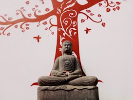 A stone statue of Buddha is seated in a meditative pose against a white wall. Behind the statue is a stylized tree painted in red, with ornate patterns, leaves, and butterflies extending from its branches.