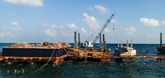 A construction platform is situated on the ocean with a large crane positioned prominently. The structure appears rusted and worn, with piles extending into the water. Concrete structures are visible on the platform, indicating ongoing construction work. The sky is clear with scattered clouds, and the water is relatively calm.