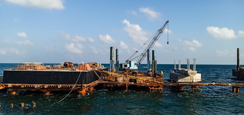 A construction platform is situated on the ocean with a large crane positioned prominently. The structure appears rusted and worn, with piles extending into the water. Concrete structures are visible on the platform, indicating ongoing construction work. The sky is clear with scattered clouds, and the water is relatively calm.