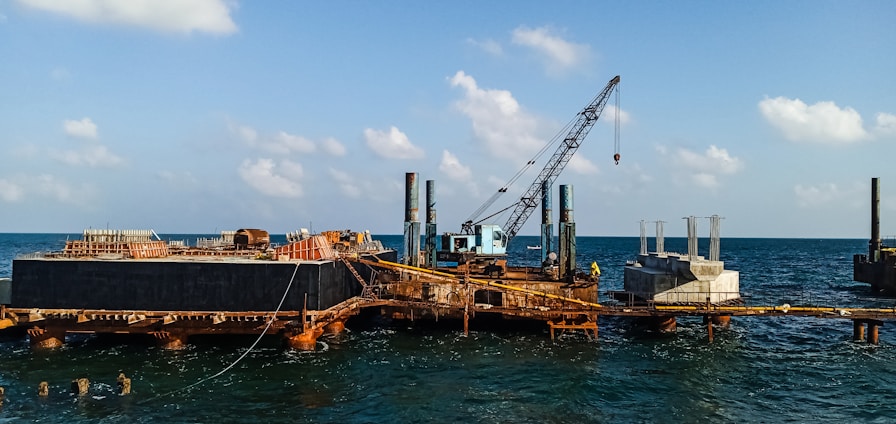 A construction platform is situated on the ocean with a large crane positioned prominently. The structure appears rusted and worn, with piles extending into the water. Concrete structures are visible on the platform, indicating ongoing construction work. The sky is clear with scattered clouds, and the water is relatively calm.