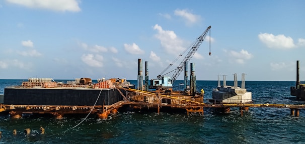 A construction platform is situated on the ocean with a large crane positioned prominently. The structure appears rusted and worn, with piles extending into the water. Concrete structures are visible on the platform, indicating ongoing construction work. The sky is clear with scattered clouds, and the water is relatively calm.