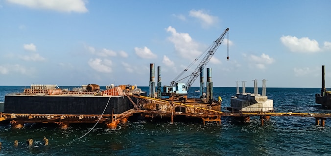 A construction platform is situated on the ocean with a large crane positioned prominently. The structure appears rusted and worn, with piles extending into the water. Concrete structures are visible on the platform, indicating ongoing construction work. The sky is clear with scattered clouds, and the water is relatively calm.