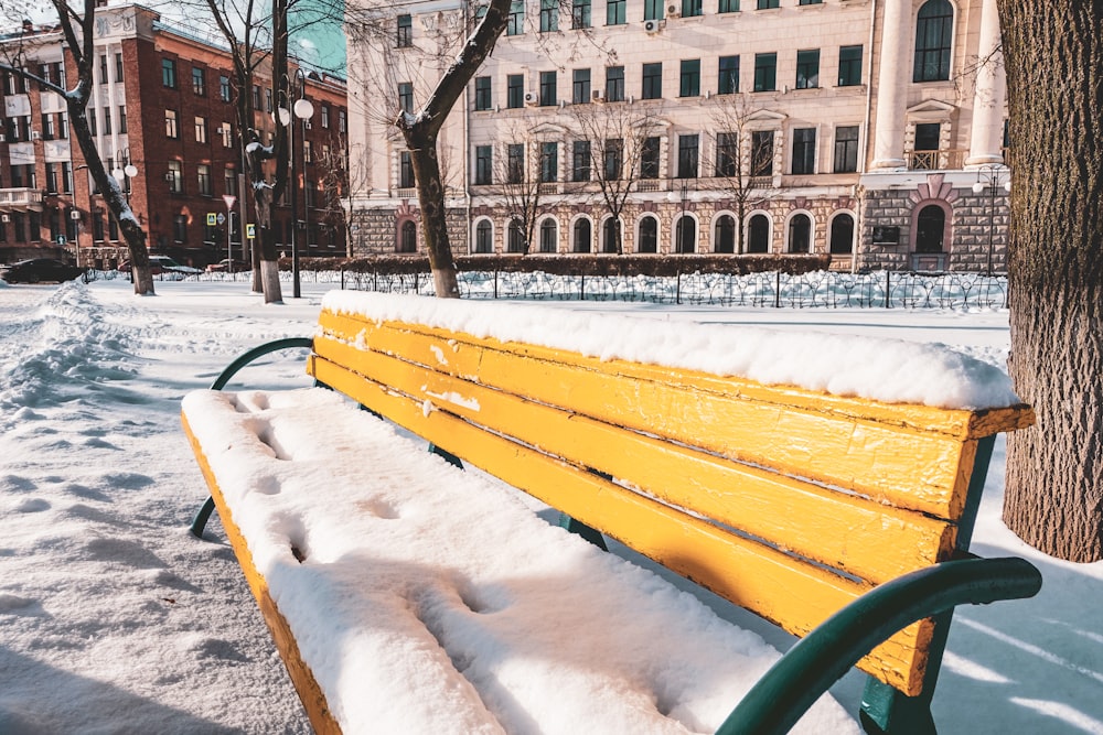 a yellow park bench covered in snow next to a tree
