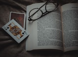 An open book lies on a textured fabric surface, with reading glasses placed on top. Beside the book, two playing cards, one featuring a jack of spades, are positioned off to the side.