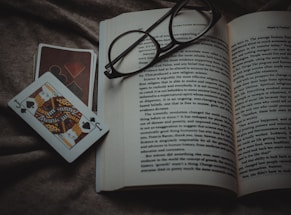 An open book lies on a textured fabric surface, with reading glasses placed on top. Beside the book, two playing cards, one featuring a jack of spades, are positioned off to the side.