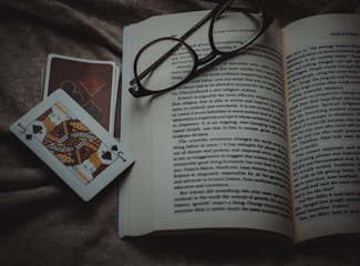 An open book lies on a textured fabric surface, with reading glasses placed on top. Beside the book, two playing cards, one featuring a jack of spades, are positioned off to the side.