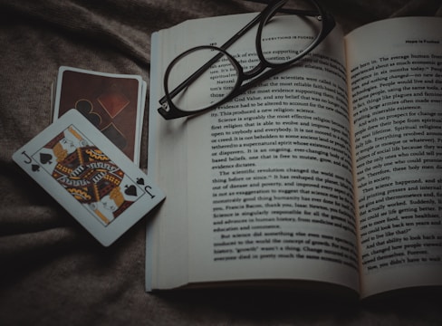 An open book lies on a textured fabric surface, with reading glasses placed on top. Beside the book, two playing cards, one featuring a jack of spades, are positioned off to the side.