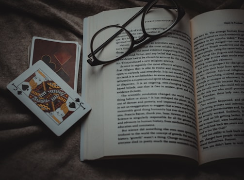 An open book lies on a textured fabric surface, with reading glasses placed on top. Beside the book, two playing cards, one featuring a jack of spades, are positioned off to the side.