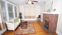 A cozy living room with hardwood floors featuring a white sofa adorned with green cushions, positioned next to large windows with beige curtains. In front of the sofa, there is a round wooden coffee table on an intricate patterned rug. A brick fireplace with decorative bottles and candle holders on the mantelpiece sits against the wall. A single tall plant is in the corner between the sofa and the fireplace.