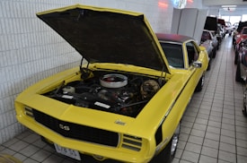 A classic yellow car with its hood open, showcasing the engine in a showroom environment. The car is surrounded by several other vehicles, all parked closely together on a tiled floor. Neon signs and white brick walls are visible in the background, contributing to a vintage or retro atmosphere.