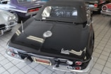 A collection of classic cars is displayed indoors on a tiled floor. The focus is on a black convertible with a distinctive Corvette logo, showcasing its sleek design and chrome accents. Surrounding the main car are other vintage cars in various colors including purple and silver.