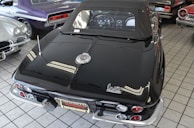 A collection of classic cars is displayed indoors on a tiled floor. The focus is on a black convertible with a distinctive Corvette logo, showcasing its sleek design and chrome accents. Surrounding the main car are other vintage cars in various colors including purple and silver.