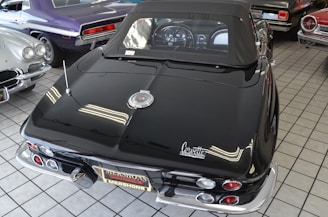 A collection of classic cars is displayed indoors on a tiled floor. The focus is on a black convertible with a distinctive Corvette logo, showcasing its sleek design and chrome accents. Surrounding the main car are other vintage cars in various colors including purple and silver.