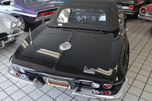A collection of classic cars is displayed indoors on a tiled floor. The focus is on a black convertible with a distinctive Corvette logo, showcasing its sleek design and chrome accents. Surrounding the main car are other vintage cars in various colors including purple and silver.