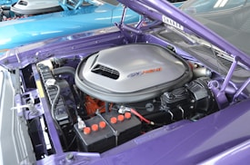 A detailed view of a car engine with a large silver air cleaner labeled '426 HEMI' on a purple vehicle. The hood is open, revealing various engine components including a battery with colored terminals, hoses, and a black engine block.