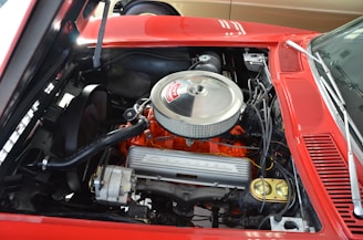 A vintage car engine with a metallic air filter cover, orange engine block, and visible wiring and components. The car is red, and various mechanical details are visible under the hood.