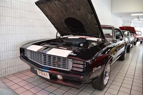 A classic muscle car with a black body and white racing stripes is parked indoors. The car has its hood raised, revealing the engine. The license plate reads 'USA-1'. The car is part of a collection lined up along a tiled floor, with other vehicles visible in the background.
