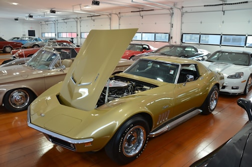A collection of vintage and modern sports cars parked inside a garage with glossy wooden flooring. The focus is on a gold-colored classic car with its hood open, revealing the engine within. Several other cars in various colors including red, blue, and white are parked nearby.