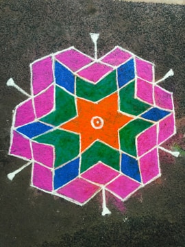 A vibrant geometric rangoli design on the ground, featuring interconnected hexagonal and star patterns in bright colors. The design includes orange, green, blue, and pink sections outlined with white lines.