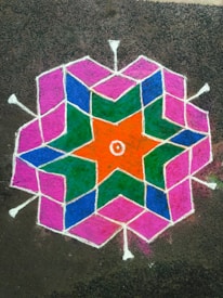 A vibrant geometric rangoli design on the ground, featuring interconnected hexagonal and star patterns in bright colors. The design includes orange, green, blue, and pink sections outlined with white lines.