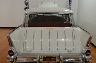 A vintage white car is shown from the rear, with chrome accents and tailfins. The car is parked indoors on a polished wooden floor with a lift in the background. The interior appears to have dark red seats.