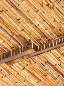 A close-up view of bamboo sticks arranged in a diagonal pattern, supported by wooden beams. The texture and natural grain of the bamboo and wood are clearly visible, showcasing earthy tones and a rustic feel.