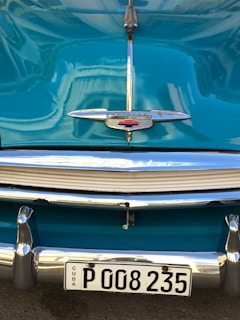 Close-up view of a vintage car's front with a shiny teal body, chrome detailing, and a license plate reading 'CUBA P 008235'. The emblem features a classic design with a wing-like shape and a red cross.
