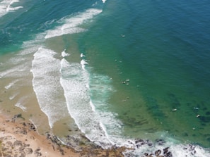 An aerial view of a coastline with waves breaking onto the shore. The turquoise and deep blue ocean transitions towards the sandy beach. Surfboards and surfers are visible in the water, waiting to catch waves.